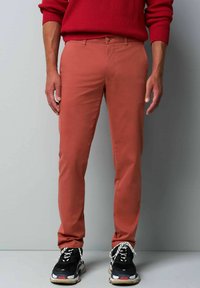 Heren slim-fit broek in gebrand oranje, met een vlakke voorkant, riemlussen en een gladde structuur, gecombineerd met zwart en beige sneakers.