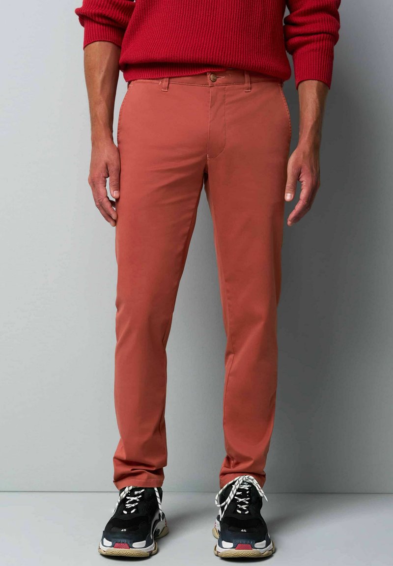 Heren slim-fit broek in gebrand oranje, met een vlakke voorkant, riemlussen en een gladde structuur, gecombineerd met zwart en beige sneakers.