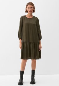 Grün kariertes Kleid mit dreiviertel Ärmeln, rundem Ausschnitt und gerafftem Rüschensaum. Getragen mit schwarzen Knöchelstiefeln.