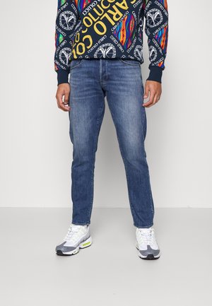 Homme portant un jean bleu, des baskets blanches et grises, et un sweat coloré avec le texte "Carlo Colucci" et des motifs graphiques.