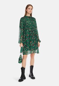 Grünes Leopardenmusterkleid mit transparenten langen Ärmeln, gerafftem Oberteil und einem lockeren Schnitt. Kombiniert mit schwarzen Kampfstiefeln und einer passenden Handtasche.