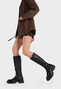 Stivali in pelle neri alti fino al ginocchio con suola spessa, cerniera laterale e finitura liscia, abbinati a una giacca marrone e pantaloni corti.
