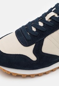 Gros plan d'une basket beige et bleu marine avec des surcouches en daim, des panneaux en tissu, des lacets foncés et une semelle en gomme.