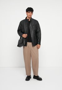 Black leather jacket with front pockets, worn over a fitted black top. Beige wide-leg trousers and black shoes complete the outfit.