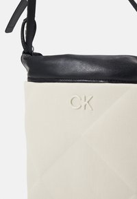 Sac à main beige et noir en tissu texturé, garniture supérieure en cuir noir, bandoulière réglable et logo "CK" en relief à l'avant.