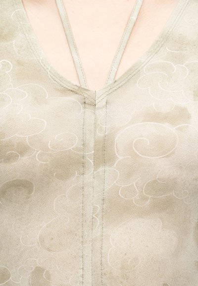 Κοντινό πλάνο σε μπεζ τοπ με λαιμόκοψη σε σχήμα V, με διακριτικά μοτίβα λευκών σύννεφων και στενούς ιμάντες ώμων που φαίνονται πάνω από το λαιμό.