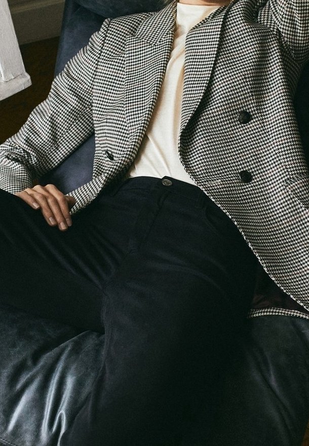Personne allongée sur un canapé en cuir foncé portant un pantalon noir, un top blanc et un blazer pied-de-poule noir et blanc à double boutonnage.