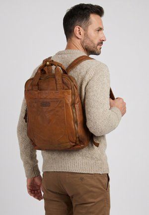 Man die een beige gebreide trui en bruine broek draagt, draagt een bruine leren Venezia-rugzak en kijkt naar rechts tegen een effen achtergrond.