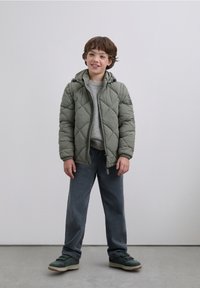 Giacca trapuntata verde imbottita con cappuccio, maglione grigio, pantaloni larghi grigi e sneaker verdi. Look casual a strati con tessuti testurizzati.