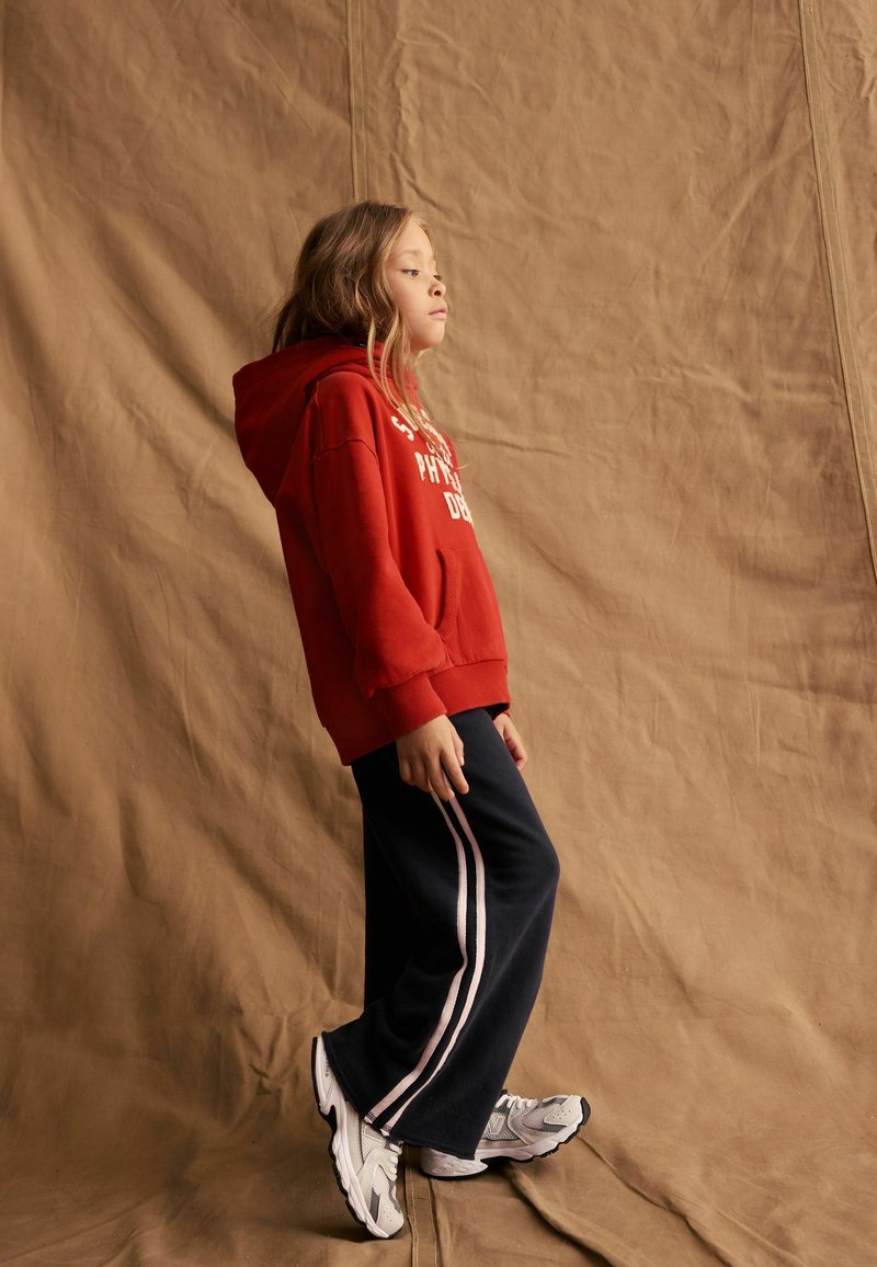 Felpa rossa con cappuccio, letteratura bianca, pantaloni ampi blu con strisce rosa e scarpe da ginnastica grigie. Sfondo semplice e texturizzato.
