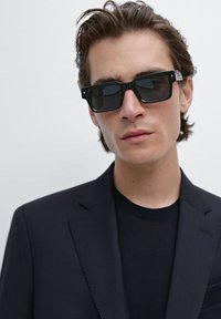 Schwarze rechteckige Sonnenbrille mit dunklen Gläsern, getragen von einer Person in einem schwarzen Rundhals-Shirt und einem gestreiften Blazer. Glatte Texturen und minimalistische Details.