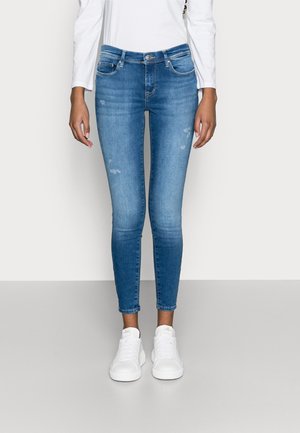 Vaqueros ajustados azul con ligero desgaste, combinados con zapatillas blancas y una camiseta blanca de manga larga, mostrados desde la mitad del torso hasta los pies.