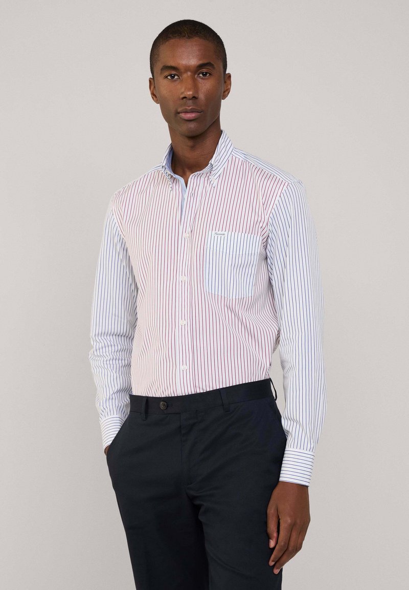 Camicia a righe con strisce verticali rosse e blu, design con colletto e taschino sul petto. Indossata con pantaloni scuri.