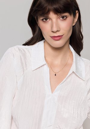 Biała, pomarszczona bluzka z kołnierzykiem i zakładką. Złoty naszyjnik z łańcuszka z małym, czarnym zawieszką, noszony na co dzień.