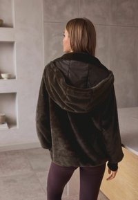 Hanorac din fleece pufoasă, de culoare maro închis, cu o glugă mare și căptușeală din satin negru. Prezintă mansete ajustate și o siluetă relaxată.