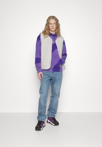 Hellgraue Steppweste über einem lila Batik-Hoodie, kombiniert mit blauen Jeans und schwarzen Turnschuhen mit lila Akzenten.