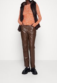 Langärmliges orangenes Oberteil mit wirbelnden Mustern, kombiniert mit braunen Lederhosen und schwarzen Plateauschuhen. Die Oberbekleidung ist eine dunkle Kunstpelzweste.