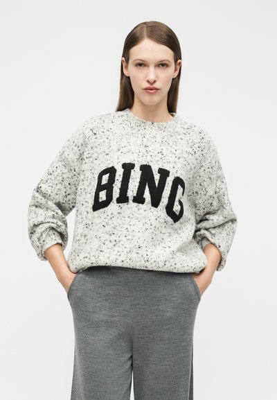 Žena s rovnými hnedými vlasmi nosí šedý sveter s farebnými škvrnami a nápisom "BING" a voľné sivé nohavice, ruky v kapsách, neutrálne pozadie.