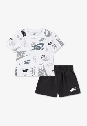 Chemise blanche à manches courtes avec des logos Nike noirs et gris partout, associée à un short noir avec un logo Nike blanc sur la jambe gauche.