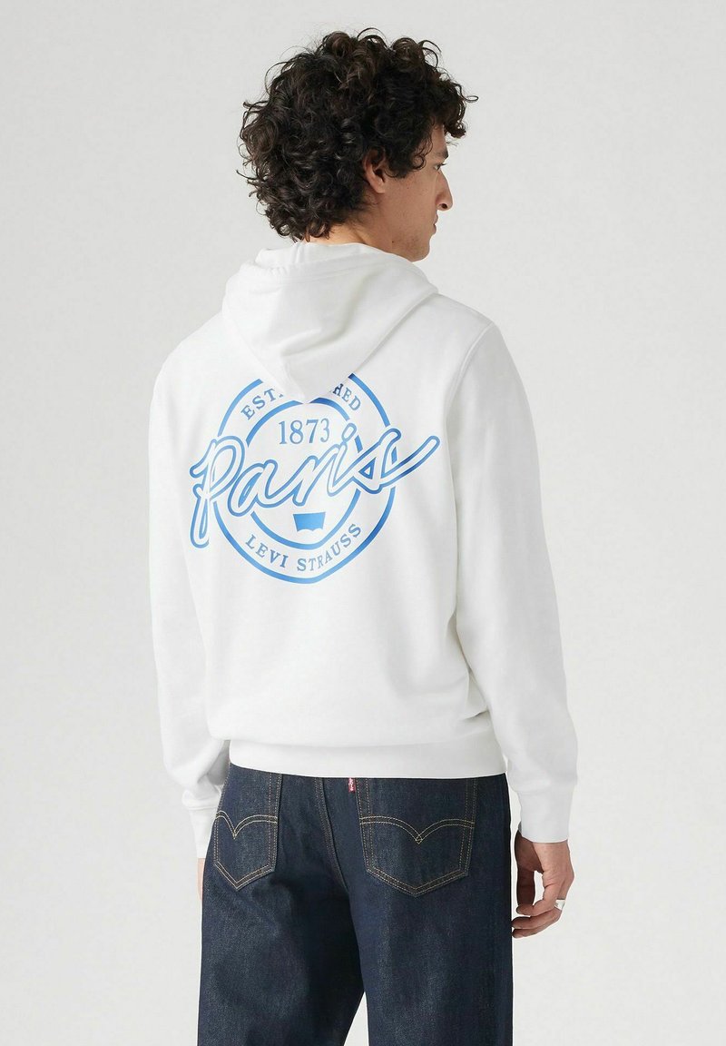 Hoodie bianco con una grafica circolare blu che mostra "Parigi" e "Fondata nel 1873". Presenta un cappuccio con cordino e polsini a costine. Indossato con jeans scuri.