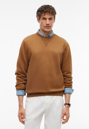 Mand iført en brun sweatshirt over en sammenfoldet denimskjorte med hvide bukser og et armbåndsur, stående mod en hvid baggrund.