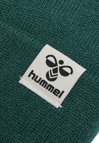 Groenblauwe gebreide stof met een wit label dat het "hummel" logo toont met een gestileerd bij-icoon. Gemedialleerde oppervlakte, close-up weergave.