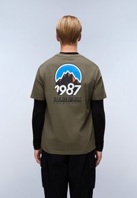 Olivově zelené tričko s grafickým designem, na kterém jsou hory, modrá obloha a na zádech nápis "1987". Krátké rukávy, volný střih.