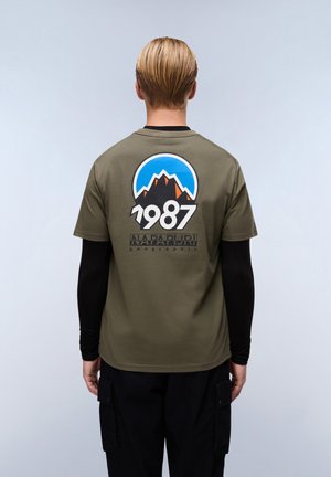 Oliven grøn t-shirt med et grafisk design, der viser bjerge, en blå himmel, og teksten "1987" på ryggen. Korte ærmer, afslappet pasform.