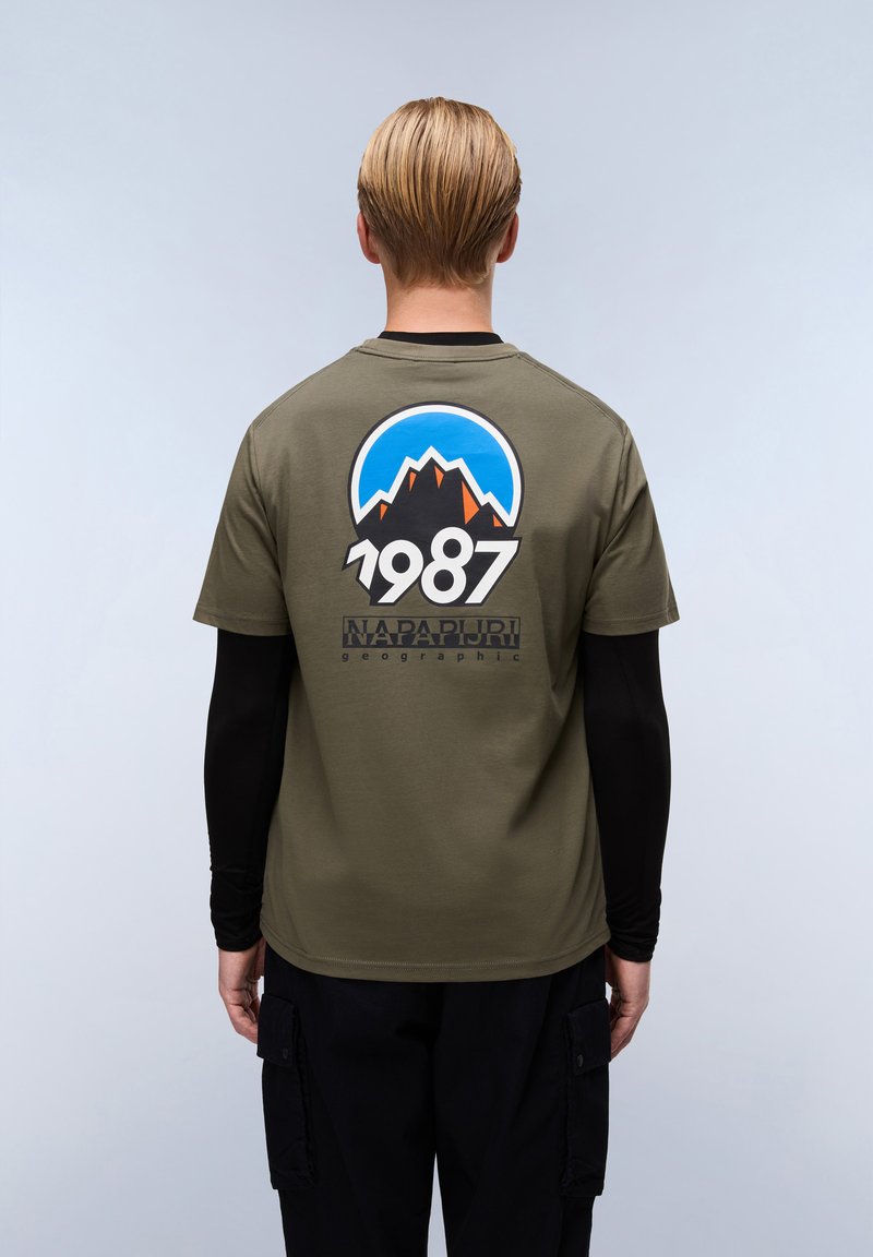 Olivově zelené tričko s grafickým designem, na kterém jsou hory, modrá obloha a na zádech nápis "1987". Krátké rukávy, volný střih.