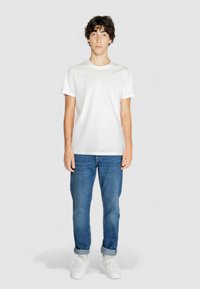 T-shirt bianca in cotone a maniche corte, con collo rotondo, indossata con jeans blu in denim e sneakers bianche, in piedi contro uno sfondo semplice.