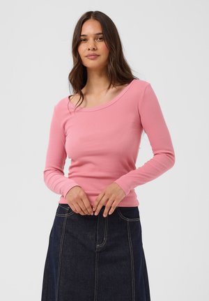 Femme aux longs cheveux bruns portant un haut rose à manches longues et une jupe en jean foncé, debout devant un fond gris uni.