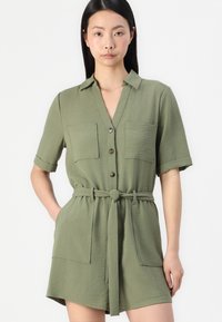 Combinaison à manches courtes de couleur vert olive en tissu texturé. Comprend un devant à boutons, deux poches poitrine et une ceinture assortie à la taille.