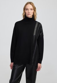 Kobieta z blond włosami, ubrana w czarny sweter z dekoltem w łódkę o teksturowanych paskach oraz czarne skórzane spodnie, na jednolitym tle.