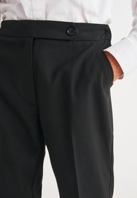 Black tailored trousers featuring a smooth texture, a button closure at the waist, and side pockets. The fabric appears durable and comfortable.