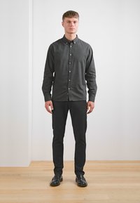 Chemise grise foncée à boutons avec un col, assortie à un pantalon noir et des chaussures noires brillantes, sur une silhouette masculine debout devant un fond uni.