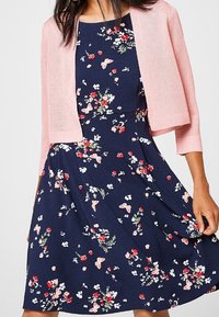 Tmavě modré květinové šaty s červenými a růžovými vzory, doplněné světle růžovým krátkým svetříkem z měkké látky s krátkými rukávy.