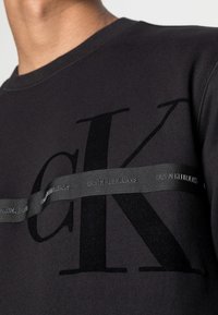 Felpa nera con un grande logo "K" nero, arricchita da un nastro grigio Calvin Klein Jeans che attraversa il petto. Tessuto morbido e testurizzato.
