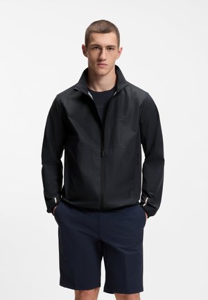 Giovane uomo con capelli corti che indossa una giacca nera con cerniera e pantaloncini blu scuro, in piedi con le mani in tasca su uno sfondo semplice.