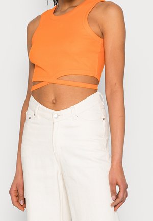 Woman wearing a sleeveless orange crop top with wrap-around straps and high-waisted white pants, shown from shoulders to hips.