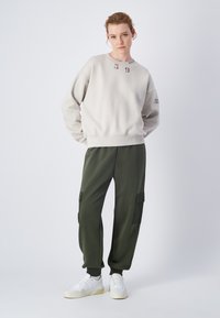 Felpa grigia chiara con colletto rotondo e grafica frontale, abbinata a pantaloni cargo verde scuro con tasche e sneakers bianche.