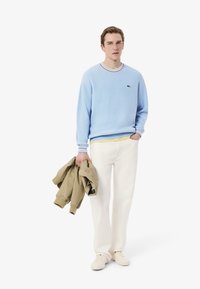 Light blue sweater with a ribbed collar featuring red, yellow, and blue stripes. Paired with white pants and beige jacket held in hand.