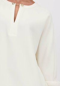 Crème lange mouwen top met een sleutelgat halslijn, vastgemaakt met gouden hardware details. Zachte stof met een gladde textuur en een losse pasvorm.