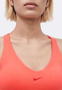 Κοντινό πλάνο ενός ατόμου που φοράει κοραλί αθλητικό σουτιέν με κόκκινο λογότυπο Nike στο στήθος, εμφανίζοντας τον λαιμό και το κάτω μέρος του προσώπου.