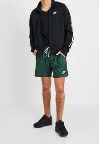 Hombre que lleva una chaqueta negra con cremallera de Nike, camiseta negra, pantalones cortos verdes oscuros de Nike con cordón blanco y zapatillas negras, de pie con las manos en los bolsillos.