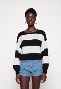 Svart og hvit stripete strikkegenser med bred utringning og ribbet nederkant, kombinert med blå denimshorts. Mykt, teksturert stoff.