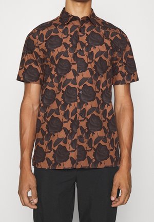 Man wearing brown short-sleeve button-up shirt with large black rose and leaf pattern, paired with black pants, against neutral background.