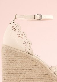 Cream wedge sandal with cut-out scalloped edge, adjustable ankle strap, and jute-wrapped heel, featuring a textured woven design.