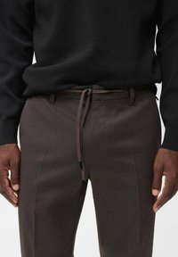 Pantaloni marroni su misura realizzati in un tessuto liscio, con vita regolabile e tasche laterali, abbinati a un maglione nero.