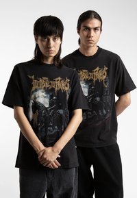 Black cotton t-shirts featuring graphic prints with gold lettering and dark imagery, short sleeves, relaxed fit, worn by two people.