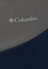 Λογότυπο Columbia τυπωμένο σε γκρι και ναυτικό μπλε ύφασμα, που δείχνει την υφή και την αντίθεση χρωμάτων του υλικού.
